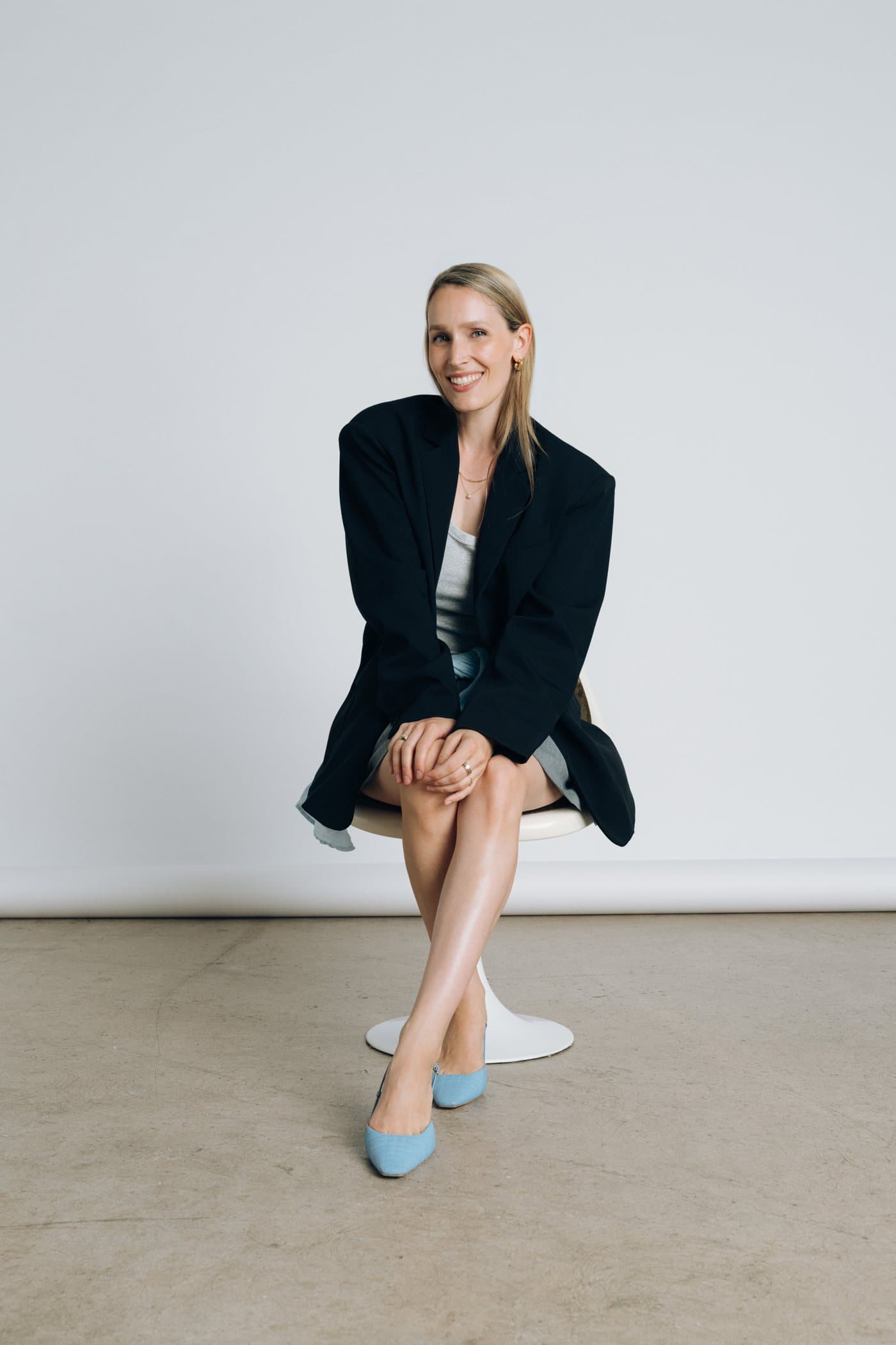 Stella Maria Sorg sitzt lächelnd auf einem weißen Stuhl, im schwarzen Blazer und mit hellblauen Schuhen